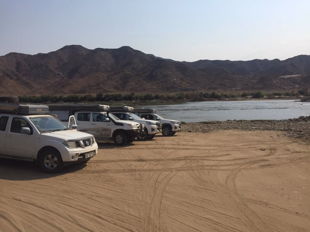 Five bakkies carrying AHA Transformers parked in a row on a sandy riverbank.